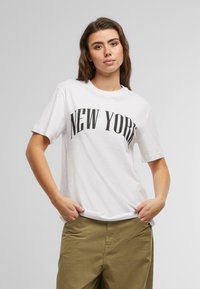 Bílá bavlněná tričko s krátkým rukávem, na kterém je velký černý text "NEW YORK." Model má na sobě olivově zelené kalhoty, ruce v kapsách, neutrální pozadí.