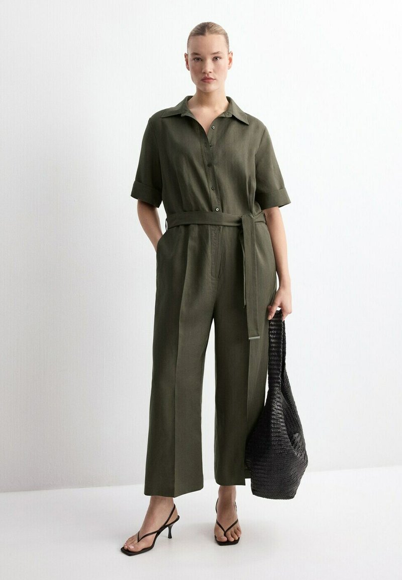 Kvinde iført olivengrøn buksedragt med bælte og korte ærmer samt sorte højhælede sandaler, og hun holder en stor sort flet taske.
