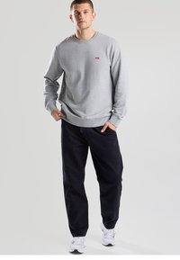 Gray sweatshirt with a round neckline and small red logo, paired with navy pants. Model wearing white sneakers. Simple, relaxed fit.