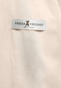 Kledinglabel op beige stof met tekst "Frieda & Freddies New York" en een klein metalen hondenembleem in het midden.