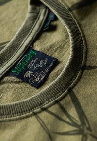 Camiseta verde oliva con un cuello redondo ribeteado, que presenta una etiqueta de marca azul oscuro. La tela tiene un patrón sutil y texturizado.
