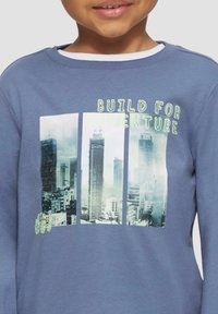 Blauwe sweatshirt met lange mouwen van katoen, voorzien van een stadslandschap grafiek en de tekst "BOUW VOOR AVONTUUR" in felgroen.