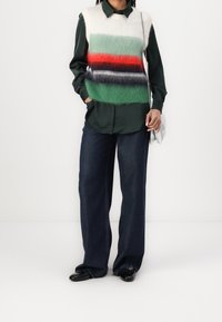 Dzianinowa kamizelka w poziome paski w odcieniach zieleni, czerwieni, czerni i bieli na ciemnozielonej koszuli z guzikami. Spodnie z szerokimi nogawkami w ciemnym denimach i czarne buty.