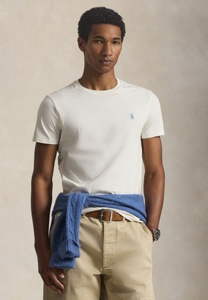 Νέος άνδρας με λευκό πουκάμισο κοντομάνικο, μπεζ παντελόνι με καφέ ζώνη, μπλε πουλόβερ δεμένο γύρω από τη μέση και ρολόι χειρός, στέκεται σε ουδέτερο φόντο.