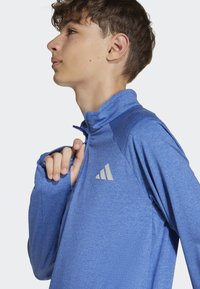 Modrý športový pulóver vyrobený z textúrovanej tkaniny, s vysokým golierom, dlhými rukávmi a strieborným logom na hrudi.