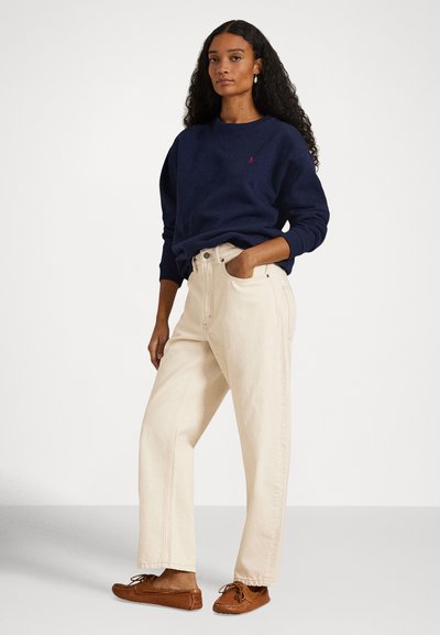 Μπλε ναυτικό φούτερ με στρογγυλή λαιμόκοψη, μπεζ τζιν φαρδιάς γραμμής με ψηλή μέση και καφέ παπούτσια σε στιλ μοκασίνια, που απεικονίζονται πάνω σε απλό φόντο.