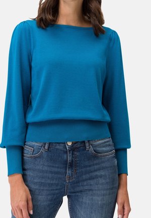 Femme portant un pull à manches longues turquoise avec des épaules bouffantes et des poignets ajustés, associé à un jean denim bleu.