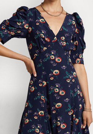 Robe fleurie en tissu bleu marine avec des manches bouffantes, un décolleté en V et une jupe évasée. Présente des motifs de fleurs multicolores et des accents de feuillage vert.