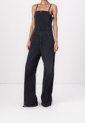 Femme portant une salopette en jean noire à jambes larges avec fines bretelles et une encolure carrée, debout devant un fond blanc uni.