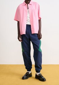 Ljusrosa kortärmad skjorta med knappar, vit linne under, marinblå träningsbyxor med gröna sidoflikar och svarta skor med spännen.