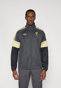 Čierno-žltá atletická bunda s vysok�ým golierom, obsahujúca logá Adidas a LFC, so zipsovým zapínaním a elastickými manžetami.