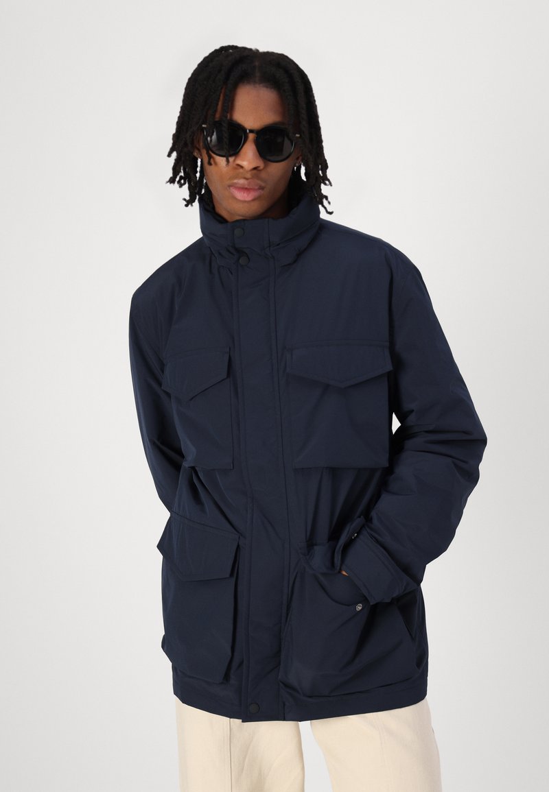 Marineblaue wasserdichte Jacke mit hohem Kragen, vier Außentaschen und Knopfverschluss. Sie verfügt über eine glatte Textur und eine lockere Passform.