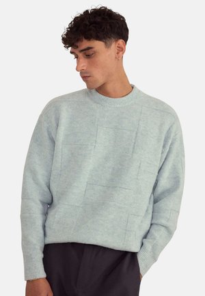 Pull bleu clair en tissu texturé, doté d'un col rond, de manches longues et d'un motif composé de sections rectangulaires.