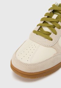 Αθλητικό παπούτσι με επάνω μέρος από κρεμ δέρμα, ελαφρώς μπεζ επενδύσεις από σουέντ, σκούρα πράσινα κορδόνια και σόλα από καουτσούκ gum, με οπές για διαπνοή.