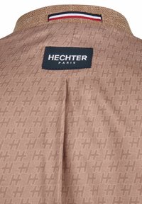 Bruine stof met een geperforeerd geometrisch patroon. Heeft een tan kleurige kraag met een geweven textuur en een zwart label met "HECHTER PARIS" erop gedrukt.