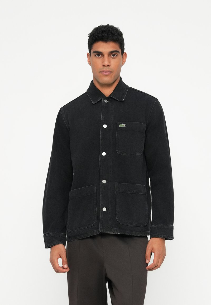 Giacca in denim nero con chiusura a bottoni, due tasche frontali e dettaglio del logo verde. Presenta un colletto e cuciture bianche a contrasto.