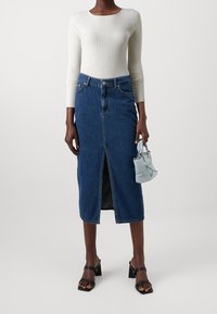 Dopasowana, sięgająca do połowy długości spódnica jeansowa w ciemnym niebieskim kolorze z przednim rozcięciem. Biały, prążkowany top z długim rękawem i mała jasnoniebieska torebka. Czarne sandały na obcasie.