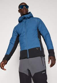 Giacca isolante blu con cappuccio, maniche nere e tasche con zip. Presenta un design elegante e materiali a contrasto.