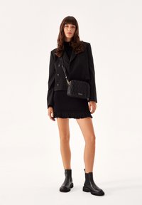 Bolso de mano negro acolchado con una correa de cadena, combinado con un vestido negro ajustado con un dobladillo de flecos. Botines negros completan el look.