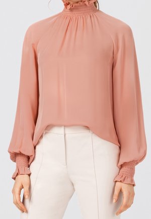 Mujer que lleva una blusa de manga larga, rosa claro, con cuello alto fruncido y puños, combinada con pantalones blancos de talle alto.