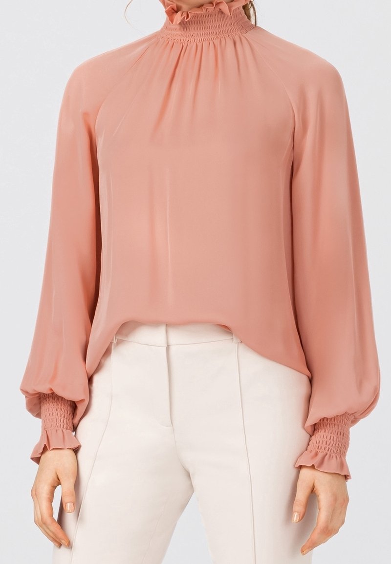 Femme portant un chemisier à manches longues rose clair avec col et poignets smockés, associé à un pantalon blanc taille haute.