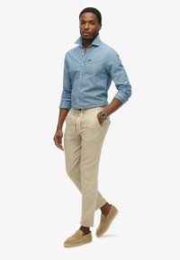 Lichtblauwe denimoverhemd met een button-down kraag, beige broek met koord, en tan instap schoenen. Horloge om de pols, staande houding.