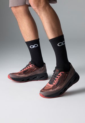 Chaussures de sport avec un motif rouge et noir, tige en mesh, semelle noire et accents rouges contrastants. Portées avec des chaussettes noires arborant un logo.