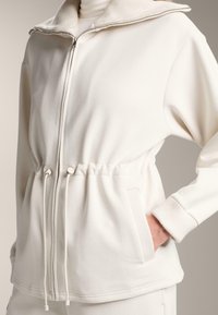 Biely zip-up sveter s rebrovaným golierom, nastaviteľným pásom na šnúrku a bočnými vreckami. Mäkká, hladká textúra.