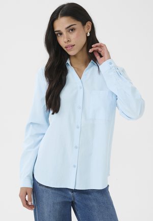Jeune femme aux longs cheveux foncés portant une chemise bleu clair boutonnée et un jean bleu, posant avec une main près du visage.