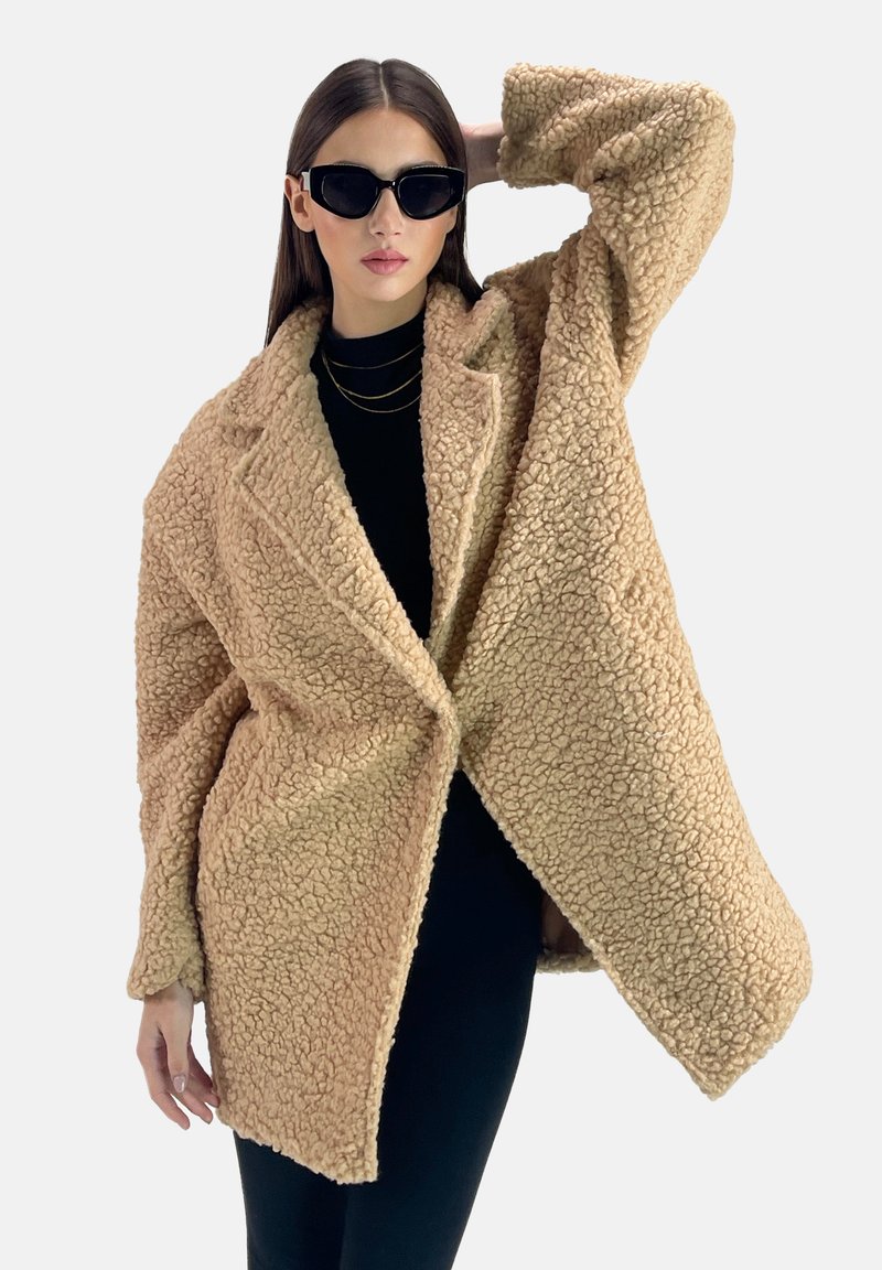 Cappotto oversized in finta pelliccia beige con un esterno testurizzato e fluffy, ampi rever e tasche frontali, indossato sopra un top nero e pantaloni aderenti.