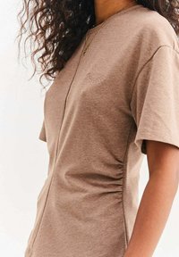 Kobieta ubrana w beżową koszulę z krótkim rękawem, z marszczeniami po bokach i małym haftem zamkniętego oka na piersi, prezentująca kręcone włosy i złoty naszyjnik.