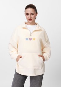 Sudadera de forro polar crema con cuello de media cremallera, bolsillo delantero y tres apliques de corazones en tonos pastel. Presenta un corte relajado y una textura suave.