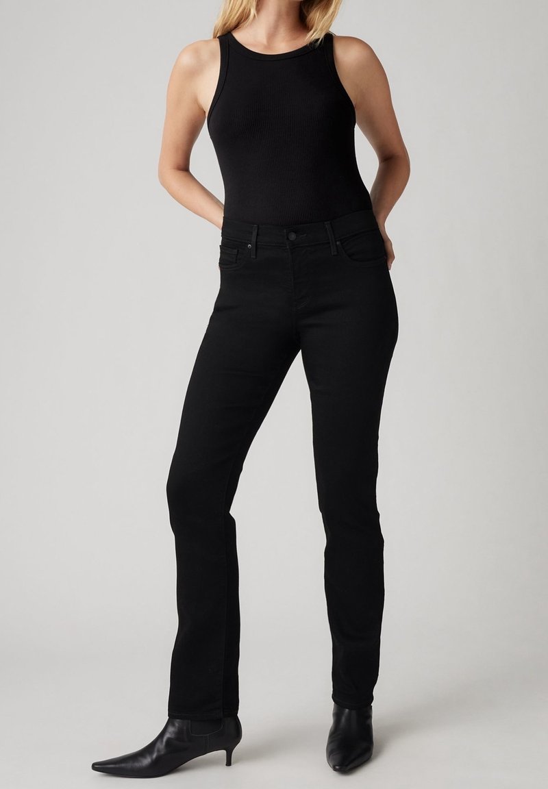 Femme portant un haut noir sans manches, un jean noir ajusté et des bottines noires à bout pointu, debout, les mains derrière le dos.