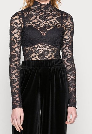 Kobieta ubrana w czarną koronkową bluzkę w kwiaty z długim rękawem i wbudowanym biustonoszem oraz czarne welurowe spodnie z wysokim stanem, stojąca na tle jednolitej ściany.