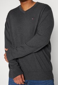 Grauer Pullover mit V-Ausschnitt aus weichem, gestricktem Stoff. Er verfügt über lange Ärmel, gerippte Bündchen und ein kleines Logo-Detail auf der Brust.