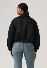 Donna con i capelli intrecciati che indossa una giacca bomber nera e jeans di jeans blu, in piedi con le spalle rivolte verso di noi su uno sfondo chiaro e semplice.