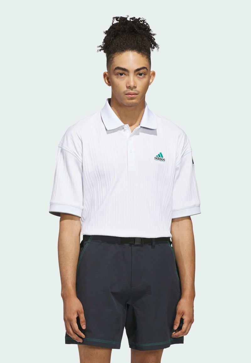 Valkoinen polo-paita, jossa on teksturoituja raitoja ja vihreä Adidas-logo, yhdistettynä tummiin shortseihin, joissa on kontrastivärinen ompelu ja musta vyö.