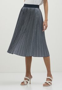 Donna che indossa una gonna midi plissettata a zigzag navy e bianca con una cintura nera e sandali con tacco bianchi e con cinturini.