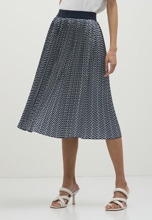 Donna che indossa una gonna midi plissettata a zigzag navy e bianca con una cintura nera e sandali con tacco bianchi e con cinturini.