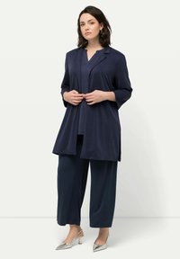 Marinblå tre-delad outfit som inkluderar en lång, öppen kappa, en topp med V-ringning och vida byxor. Strukturerad tyg med silverfärgade klackskor.