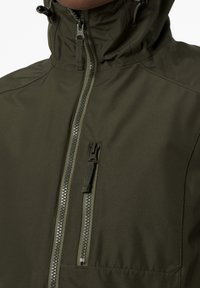 Veste vert olive avec un col montant, une fermeture éclair centrale à l'avant et une poche poitrine zippée avec une finition texturée. Matériau léger.