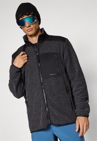 Sort og grå zip-up fleecejakke med en teksturert overflate, med glidelås-lommer og et logo-patch på brystet. Blå bukser og svart lue.
