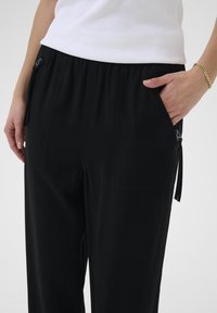 Pantaloni neri a vestibilità rilassata con vita elastica, tasche laterali e dettagli con zip. Tessuto dalla texture liscia e design minimalista.