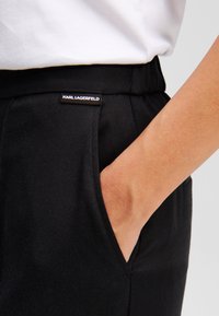 Zwarte, gestructureerde broek met een zijzak en een zichtbaar label op de tailleband met de tekst "KARL LAGERFELD." Draagwijdte op maat.