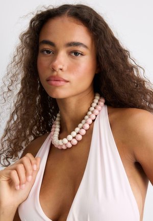 Jeune femme aux cheveux bouclés portant un collier à double rangée de perles roses et blanches et un haut sans manches rose clair, regardant droit devant.