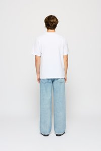 Man met kort bruin haar staat met zijn rug naar de camera, gekleed in een eenvoudig wit T-shirt, lichtblauwe wijde jeans en zwarte schoenen, tegen een witte achtergrond.