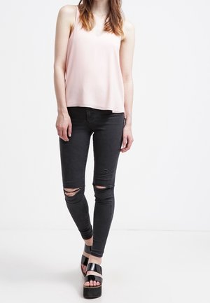 Vrouw draagt een lichtroze mouwloze top, gescheurde zwarte skinny jeans en zwarte platformsandalen, staand tegen een witte achtergrond.