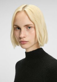 Schwarzer Pullover mit hohem Kragen und strukturiertem Strick; das Modell hat kurzes, glattes blondes Haar und einen neutralen Hintergrund, der das Kleidungsstück hervorhebt.
