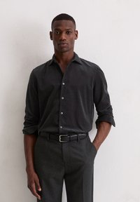 Homme portant une chemise sombre à boutons avec les manches retroussées, un pantalon sombre et une ceinture noire, se tenant contre un mur blanc avec une main dans la poche.