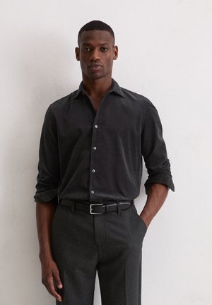 Man wearing dark button-up shirt with sleeves rolled up, dark trousers, and black belt standing against a white wall with one hand in pocket.
